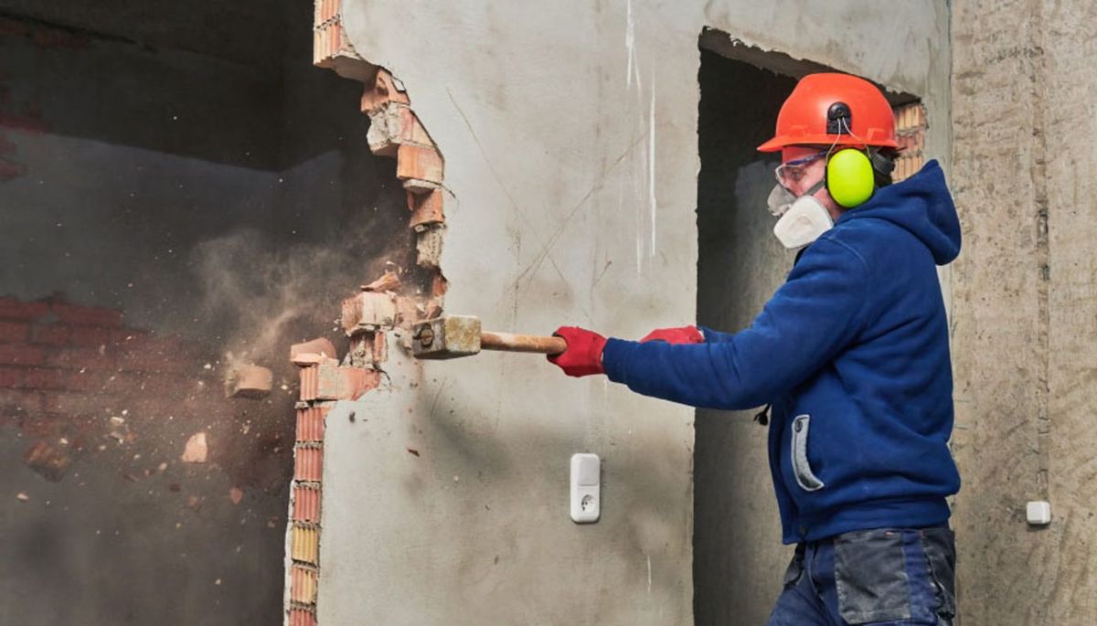 Worker with helmet, ear defenders and respirator knocking down a brick wall with a sledgehammer (illustrative image)